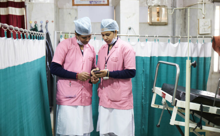 Two women in clinical scrubs look at a phone screen in a clinic.