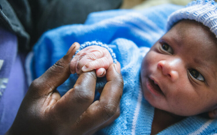 A newborn baby in a blue hat looks off camera while holding a hand. Both are of African descent.