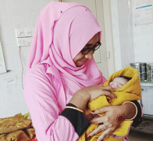 A woman in a pink hijab holds a baby swaddled in yellow.